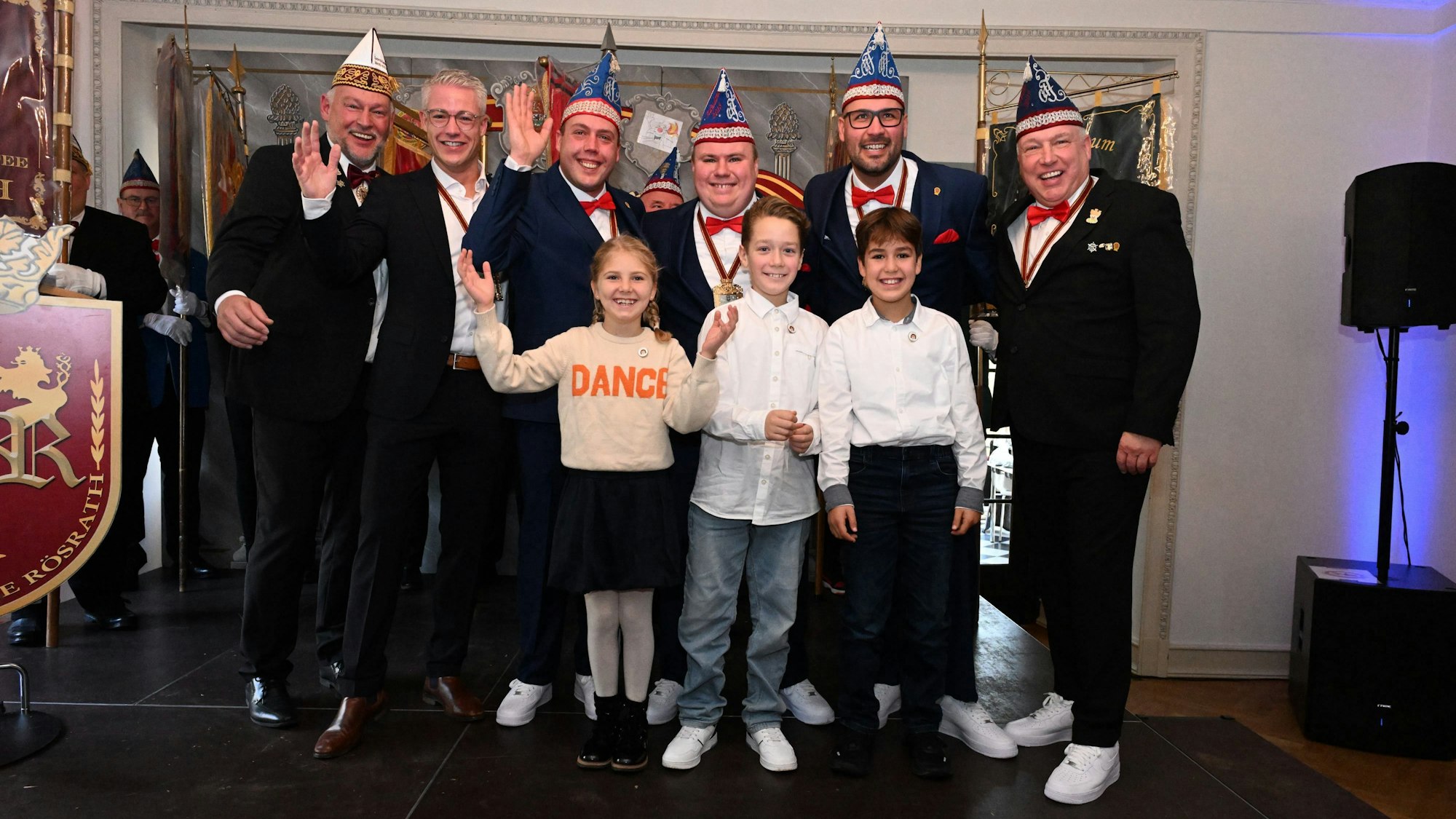 Eine Reihe von Männern mit Schiffchen auf dem Kopf steht auf einer Bühne. Vor ihnen stehen drei Kinder, zwei Jungen und ein Mädchen.