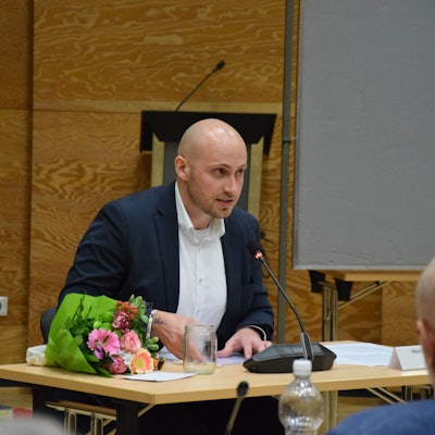 Ein junger Mann mit Glatze sitzt an einem Schreibtisch und spricht in ein Mikrofon.