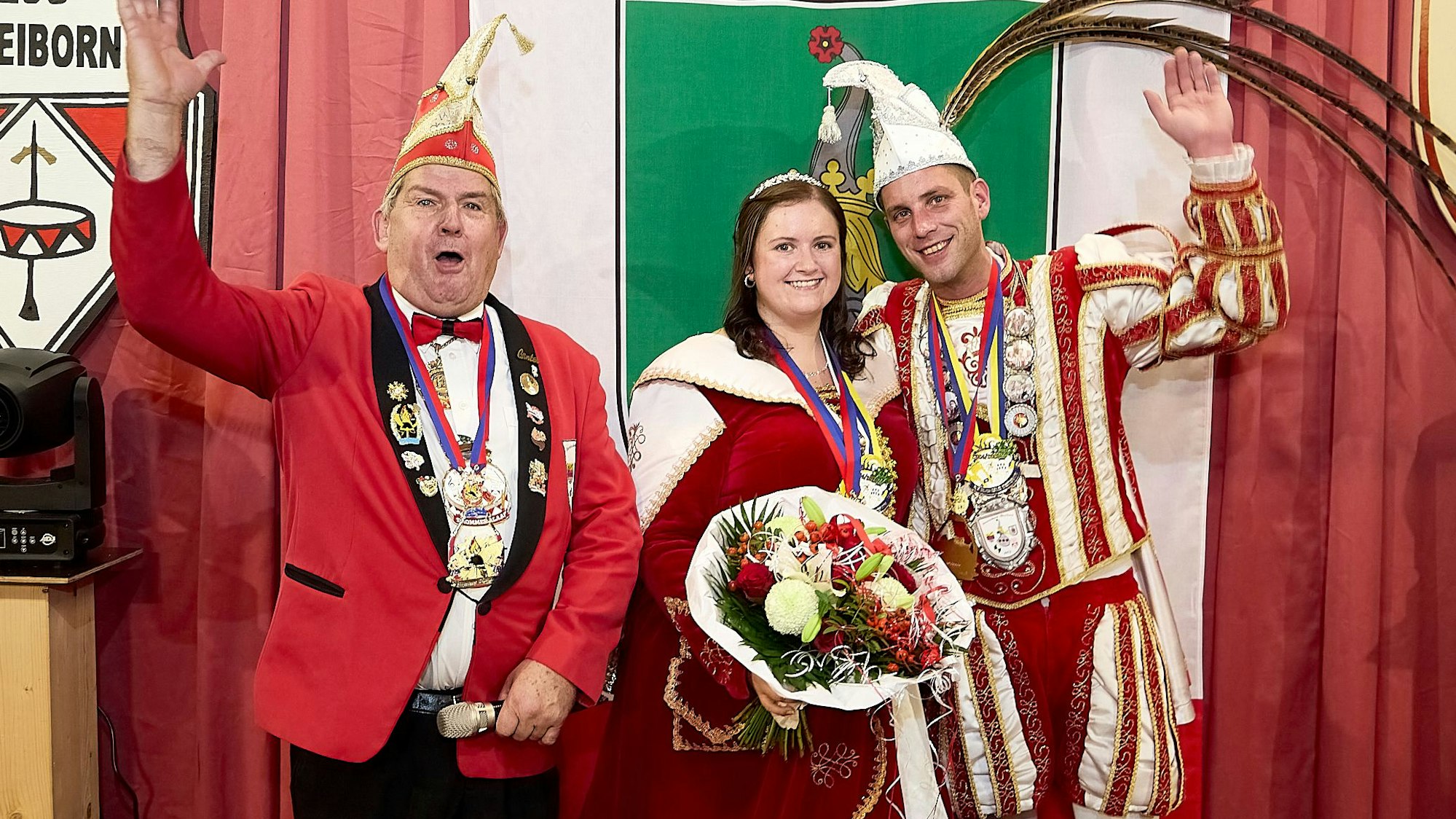 Vor einem grün-rot-weißen Vorhang stehen das Prinzenpaar aus Dreiborn im Ornat und der Sitzungspräsident. Die Männer winken.