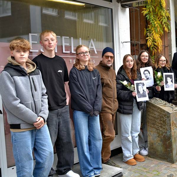 Am Gedenkstein der einstigen Synagoge in Bad Münstereifel stehen einige Menschen. Manche halten Schwarz-Weiß-Fotos oder weiße Rosen in den Händen.