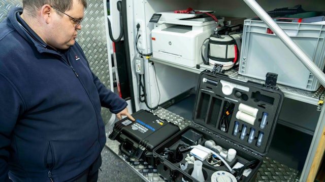 Ein Mann in einer blauen Jacke steht an der geöffneten Hecktür eines Fahrzeugs, in dem mehrere Kisten verlastet sind. Ein Koffer mit technischer Ausrüstung ist geöffnet.