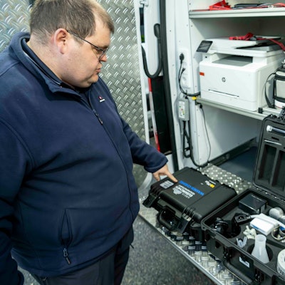 Ein Mann in einer blauen Jacke steht an der geöffneten Hecktür eines Fahrzeugs, in dem mehrere Kisten verlastet sind. Ein Koffer mit technischer Ausrüstung ist geöffnet.