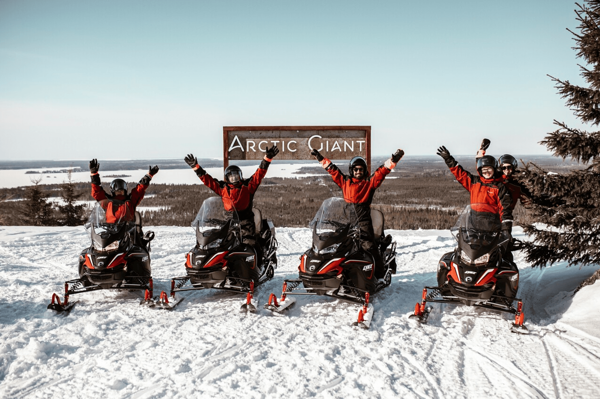 Eine Schneemobil-Tour in Finnland. 4 Schneemobile, darauf Menschen die in die Kamera lächeln und die Arme in die Luft strecken.