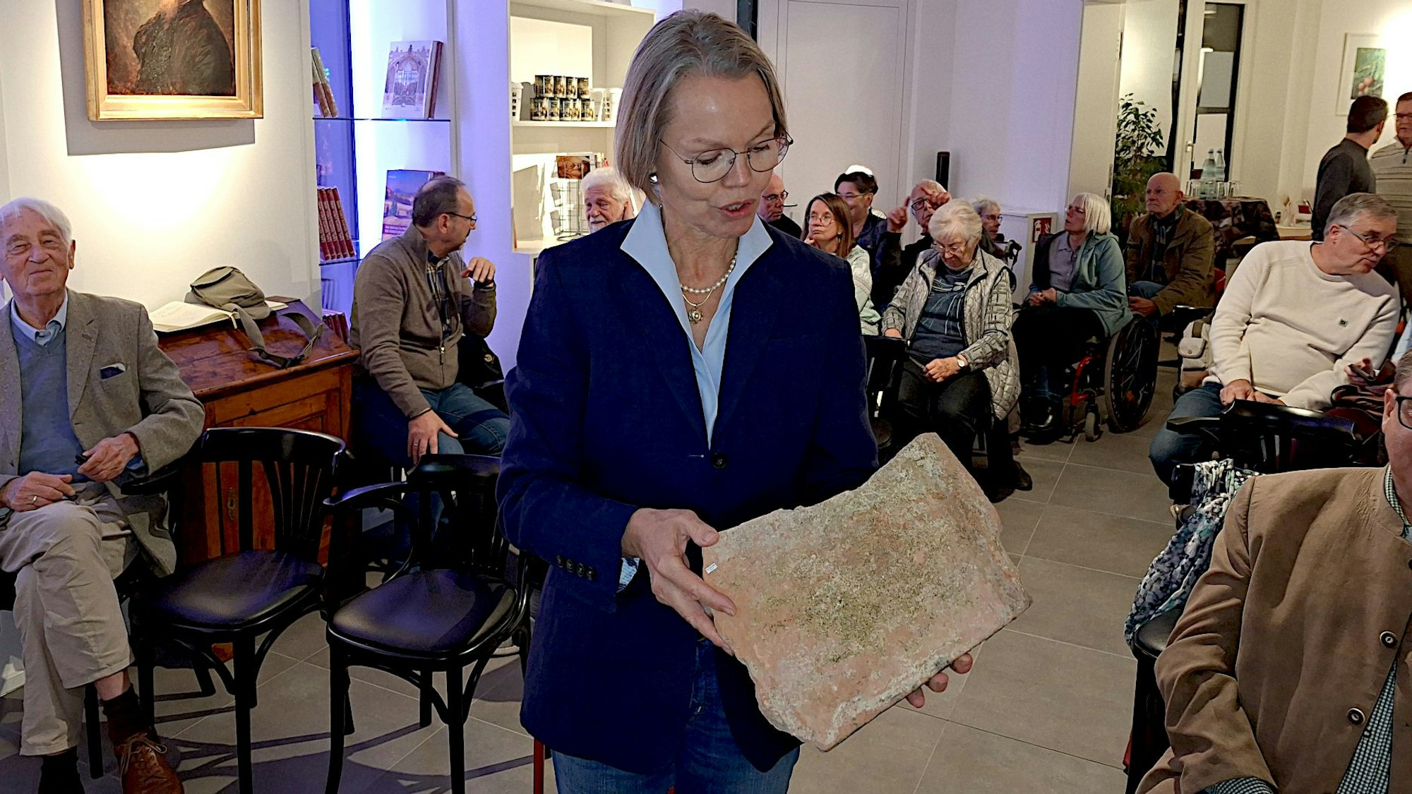 LVR-Archäologin Dr. Ulrike Müssemeier hält einen römischen Ziegel in den Händen. Hinter und neben ihr sitzt das Publikum.