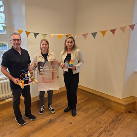 Drei Personen, eine hält ein Plakat, die anderen Spielzeug.