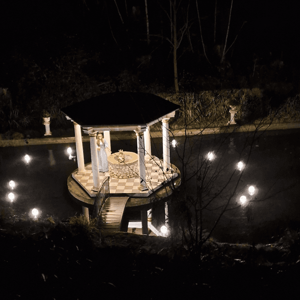 Ein Teich im Dunkeln. Lichter beleuchten eine kleine Insel mit einem Pavillon. Hier sitzt auf dem Brunnenrand der Froschkönig, daneben eine Figur, die aussieht wie eine Prinzessin