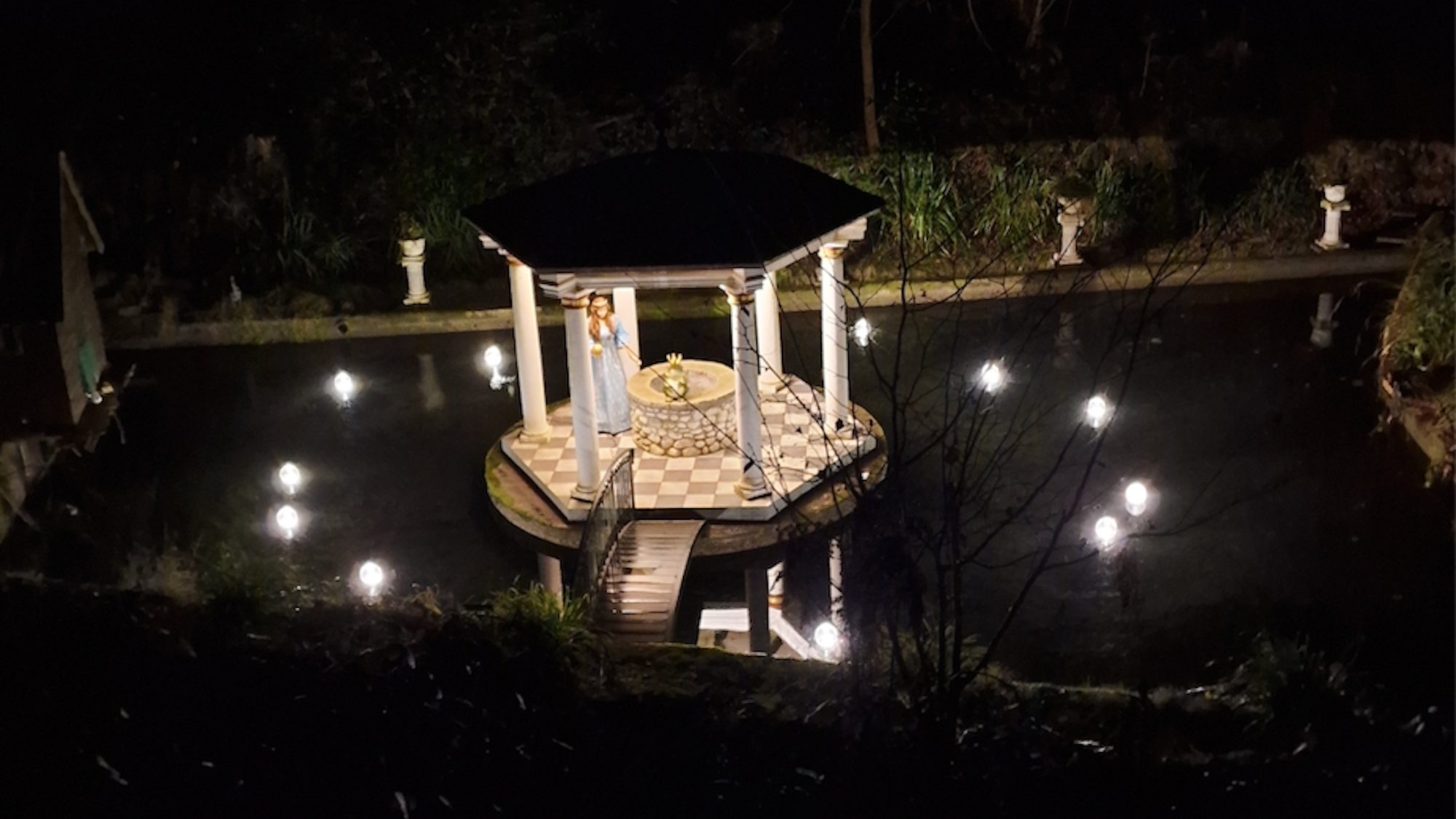 Ein Teich im Dunkeln. Lichter beleuchten eine kleine Insel mit einem Pavillon. Hier sitzt auf dem Brunnenrand der Froschkönig, daneben eine Figur, die aussieht wie eine Prinzessin