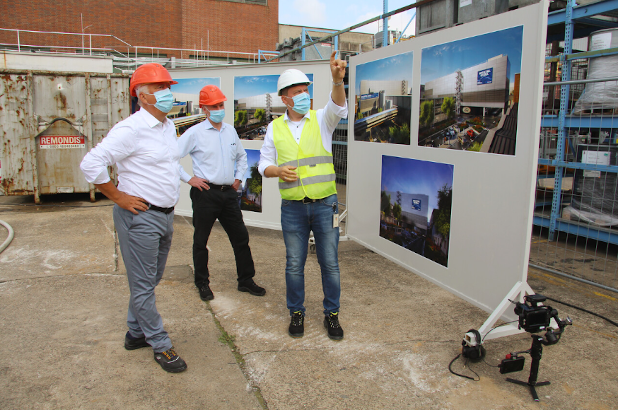 Drei Männer stehen mit Helmen auf einer Baustelle und schon sich auf einer Tafel Bilder eines Umgestaltungs-Projekts an.