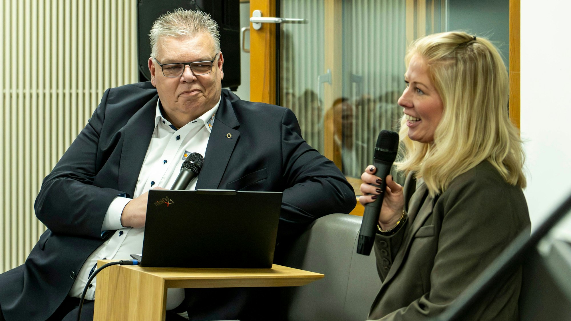 Jochen Kupp und Sarah Komp sitzen auf einer Bank und halten Mikrofone in den Händen.