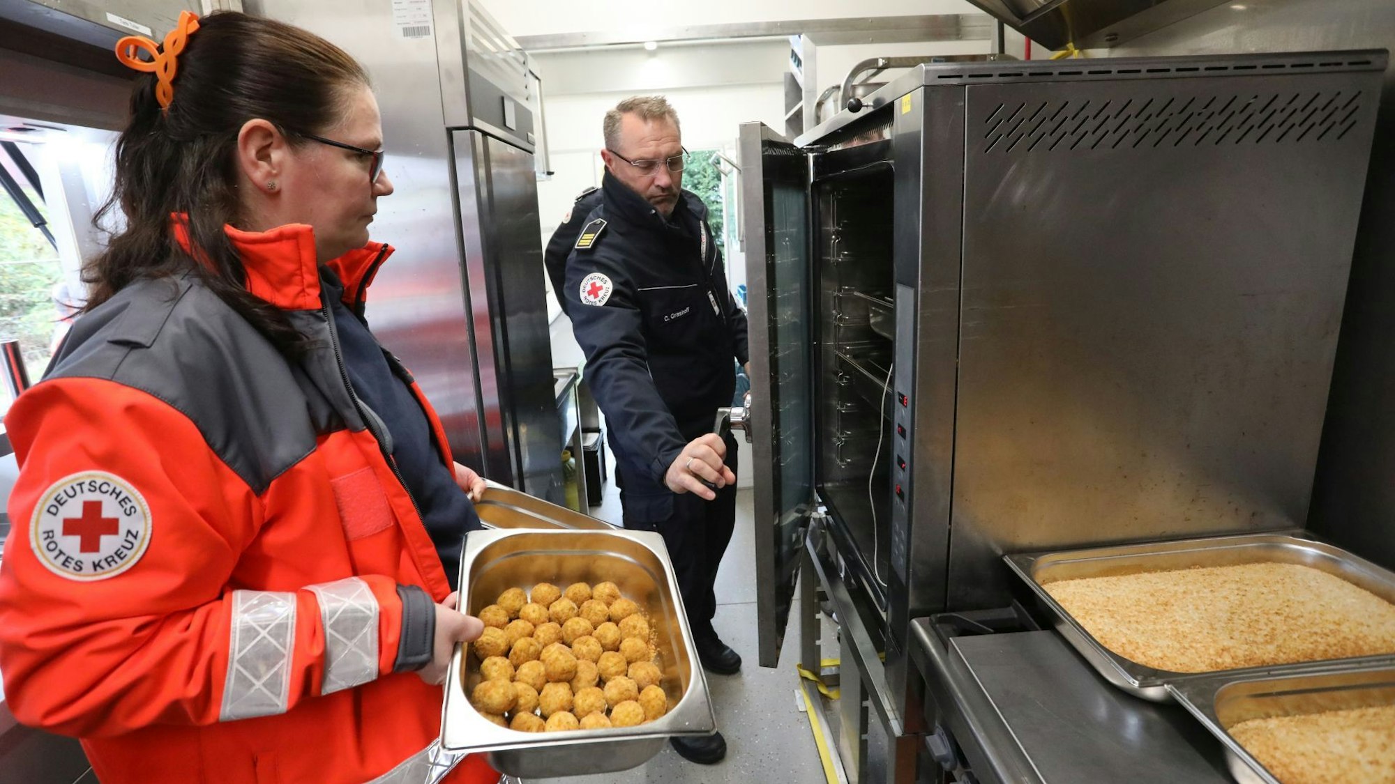 Ein Mann und eine Frau an Küchengeräten, sie trägt eine rote Jacke mit DRK-Emblem.