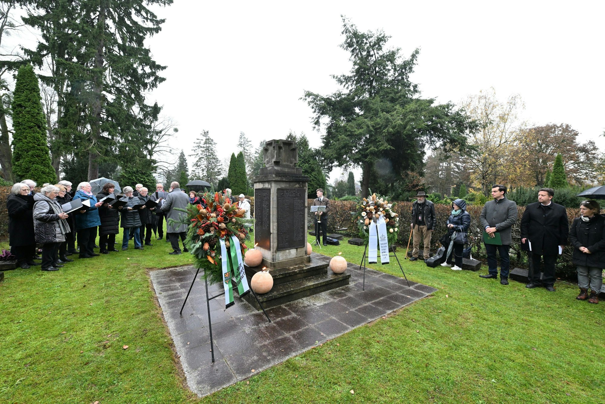 Eine Trauergemeinde hat sich um ein Ehrenmal mit Trauerkränzen versammelt. Die Besucher tragen dunkle Kleidung, einige halten Liedtexte in der Hand.