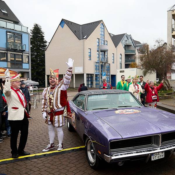 Der Prinz steht im Ornat vor dem violetten amerikanischen Schlitten und reckt die Hand zum Jubel hoch.
