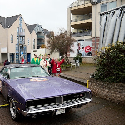 Der Prinz steht im Ornat vor dem violetten amerikanischen Schlitten und reckt die Hand zum Jubel hoch.