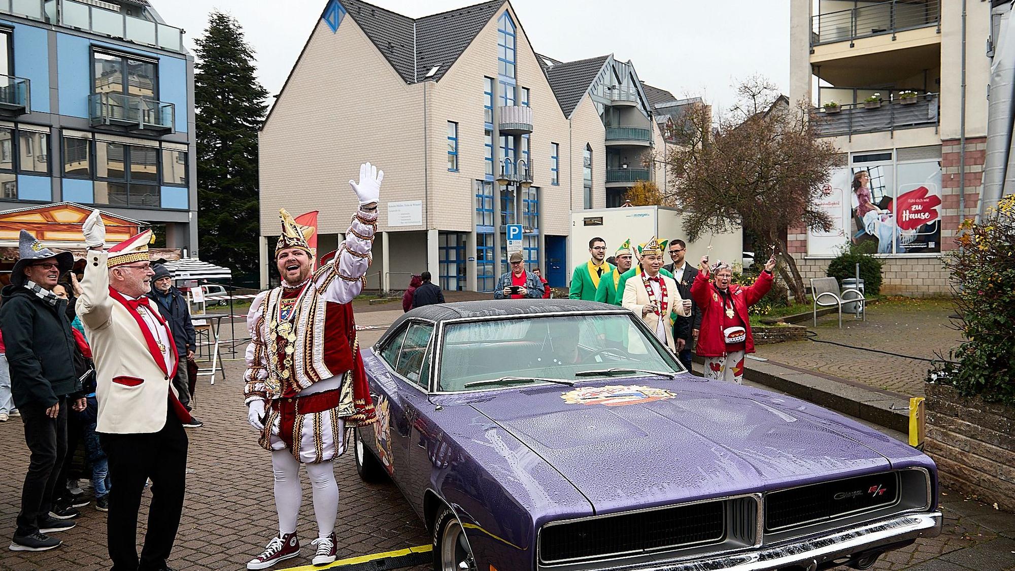 Der Prinz steht im Ornat vor dem violetten amerikanischen Schlitten und reckt die Hand zum Jubel hoch.