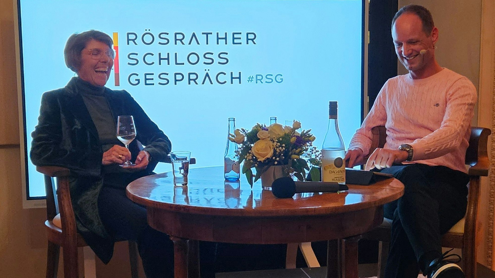 Eine Frau mit einem Glas Wein in der Hand sitzt an einem Tisch neben einem Mann. Beide lachen.