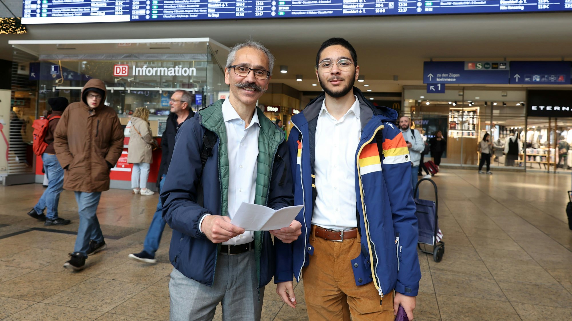 Zwei Männer schauen in die Kamera. Sie stehen in der Ankunftshalle des Kölner Hauptbahnhofs.