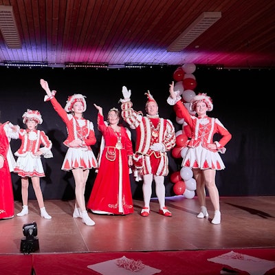 Auf der Bühne stehen die Jugendprinzessin und das Prinzenpaar mit Gardetänzerinnen und heben ihre Arme zum Gruß an die Narren.