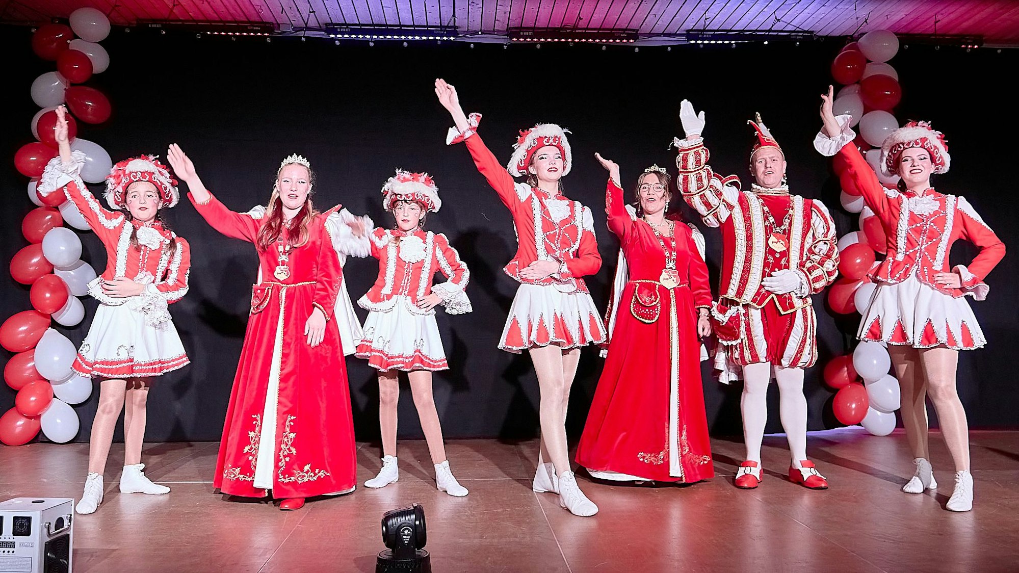 Auf der Bühne stehen die Jugendprinzessin und das Prinzenpaar mit Gardetänzerinnen und heben ihre Arme zum Gruß an die Narren.