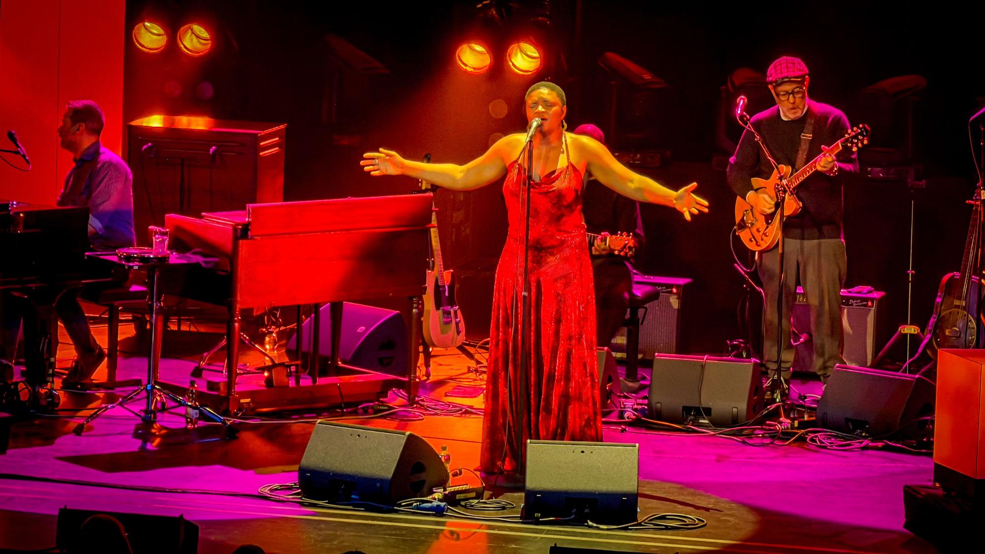 Die Sängerin Lizz Wright steht auf der Bühne hinter einem Mikrofonständer, sie trägt ein rotes Abendkleid und hat ihre Hände weit geöffnet zu einer einladenden Geste. Links hinter ihr ist ein Musiker am Piano zu erkennen, rechts ein Gitarrist.