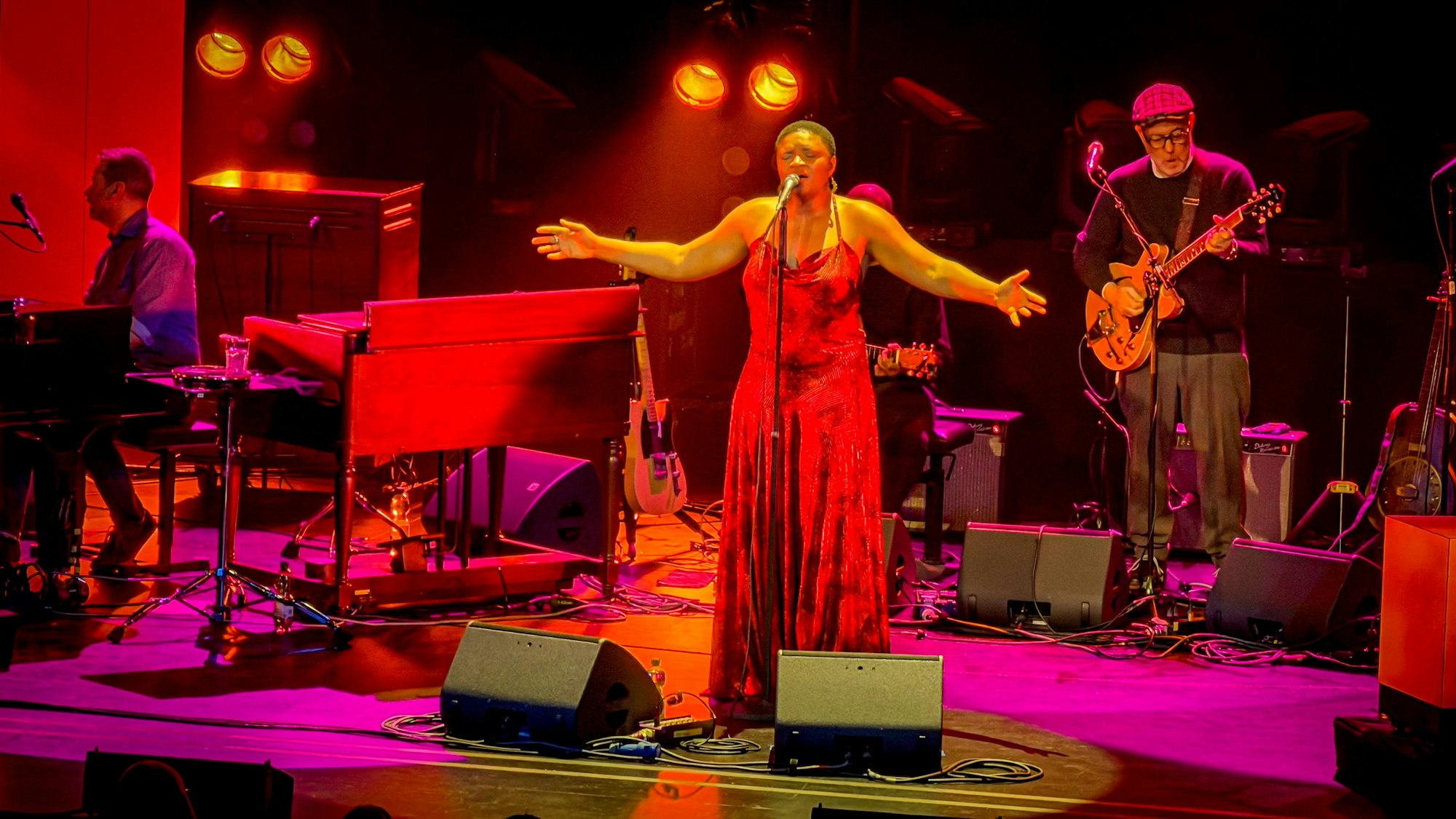Die Sängerin Lizz Wright steht auf der Bühne hinter einem Mikrofonständer, sie trägt ein rotes Abendkleid und hat ihre Hände weit geöffnet zu einer einladenden Geste. Links hinter ihr ist ein Musiker am Piano zu erkennen, rechts ein Gitarrist.