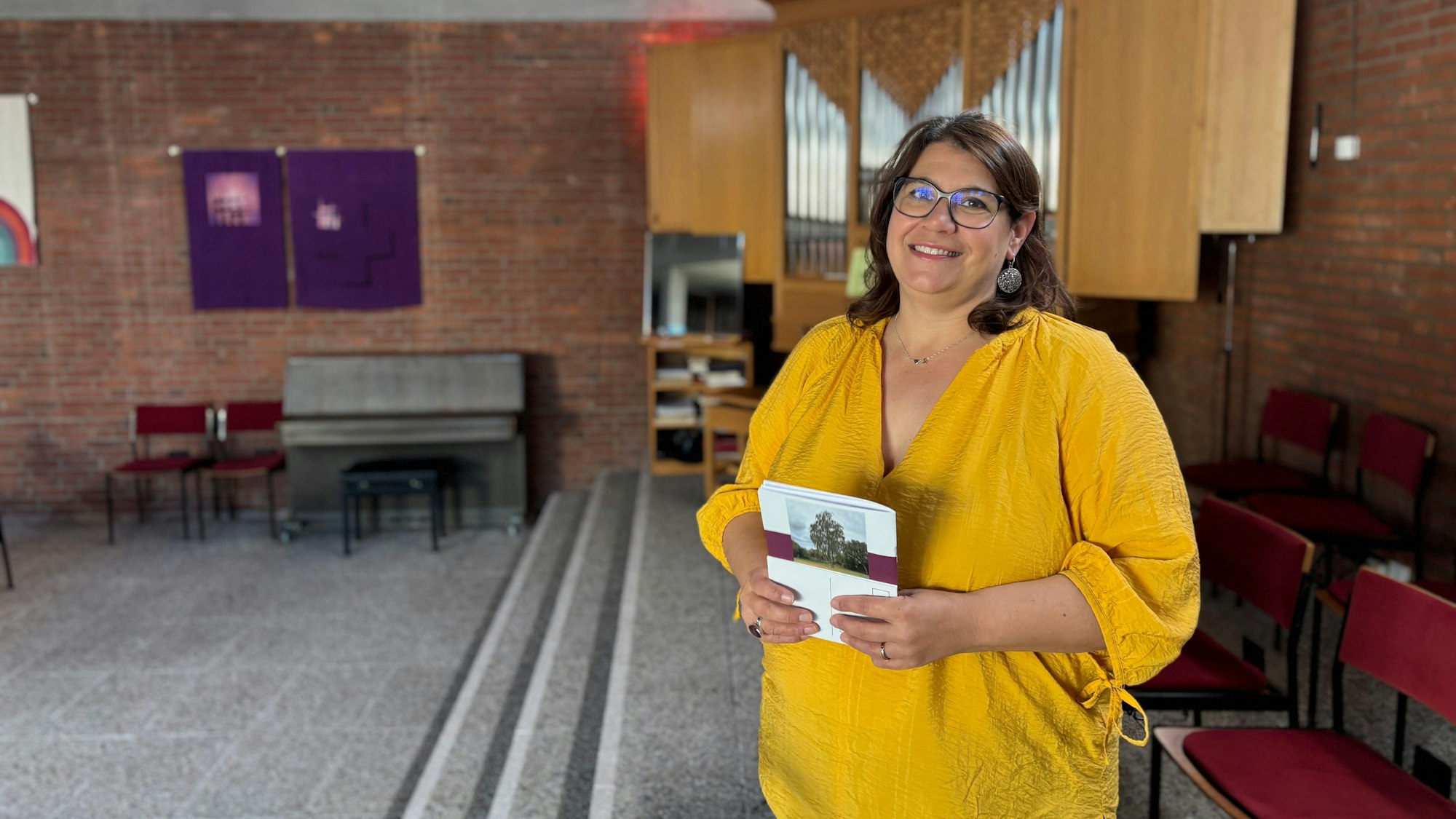 Judith Weichsel hält die Zeitung der Evangelischen Kirchengemeinde Weilerswist vor sich. Sie steht in einem Raum der Martin-Luther-Kirche, in dem sich eine Orgel befindet.