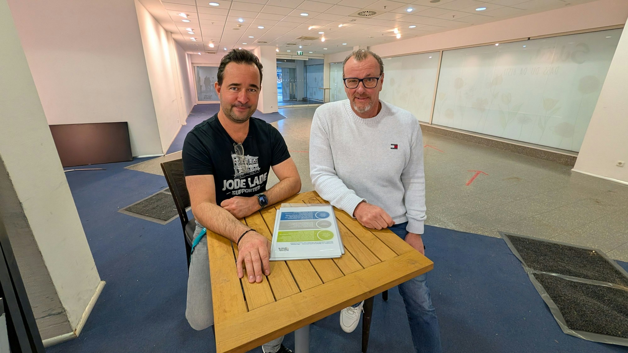 Das Foto zeigt die beiden Betreiber auf Hockern an einem Stehtisch in den Räumen der früheren Postfiliale im Hürth-Park.