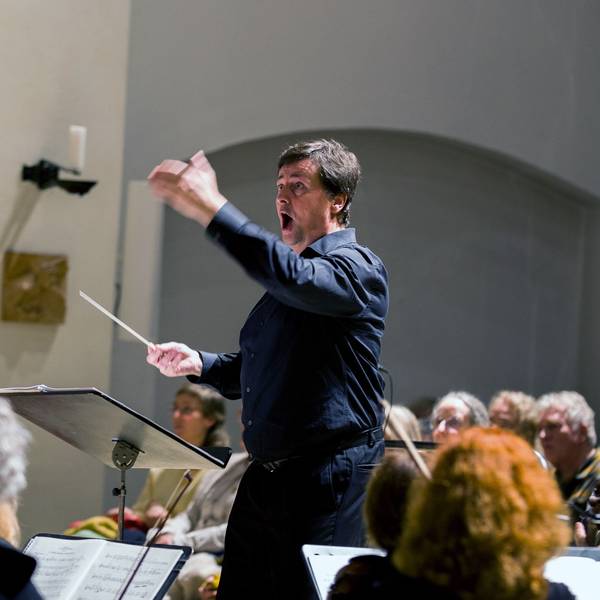 Ein Mann in schwarzem Hemd und schwarzer Hose dirigiert Chor und Orchester.