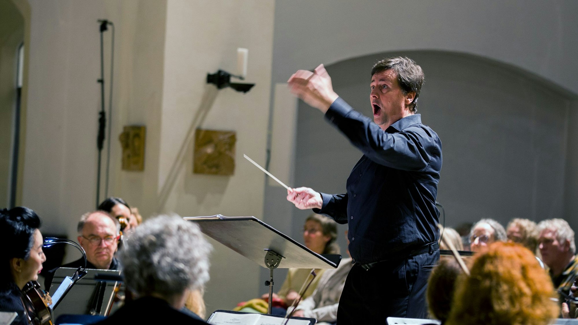 Ein Mann in schwarzem Hemd und schwarzer Hose dirigiert Chor und Orchester.