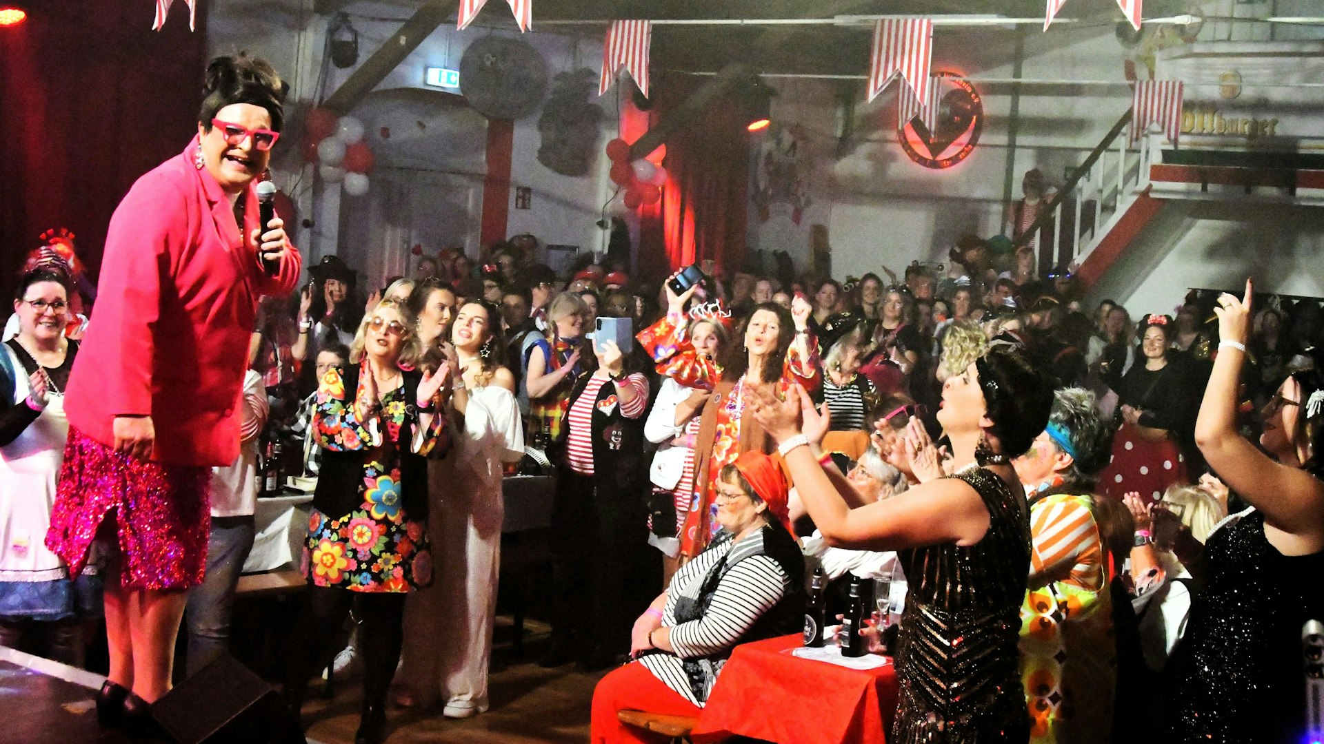 Lieselotte Lotterlappen steht mit Mikrofon in der Hand vor den jubelnden Jecken.