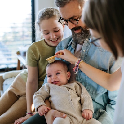 Eine junge Familie sitzt zusammen mit einem Baby und einer Erziehungshelferin auf dem Sofa. Sie kämmt das kleine Kind.