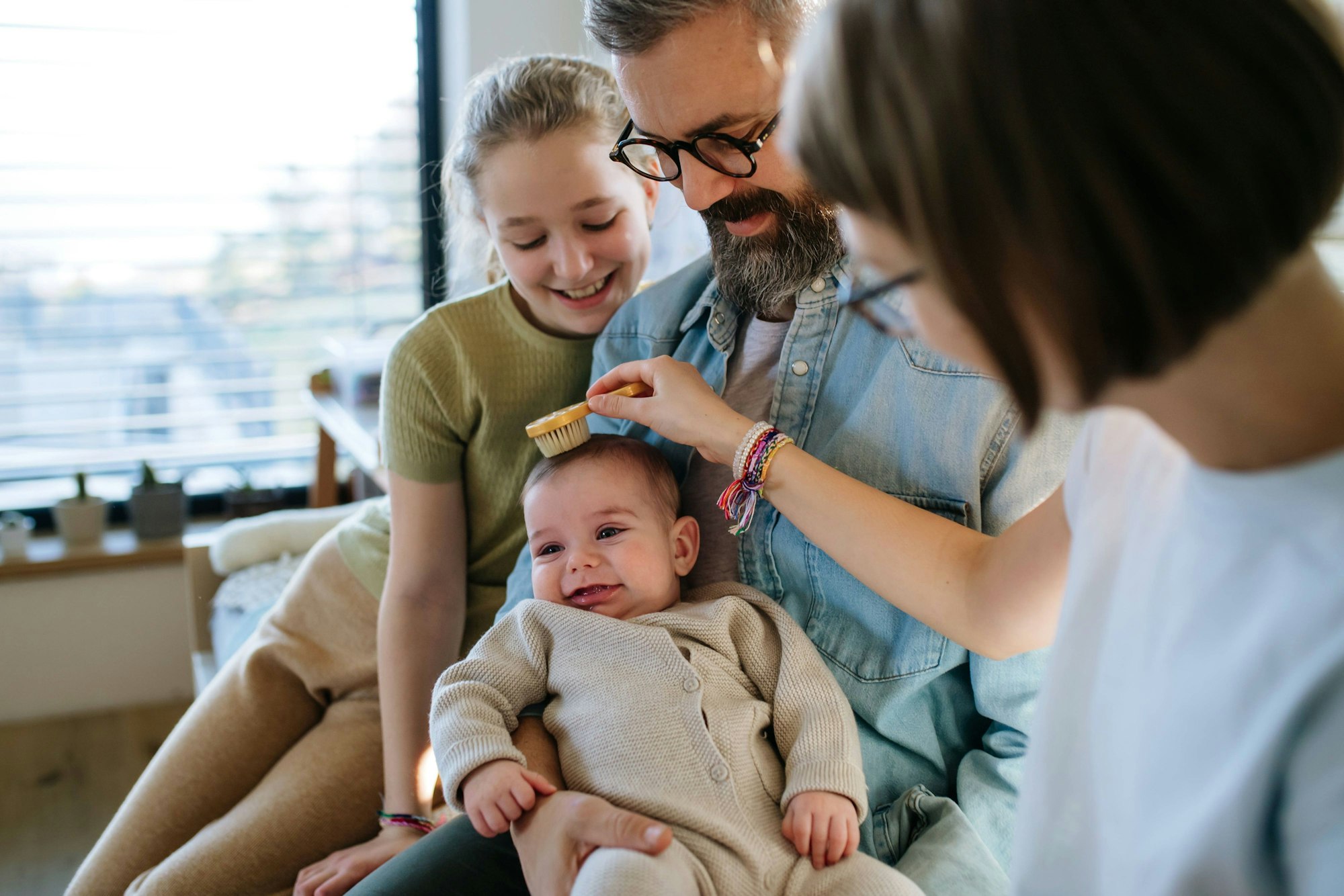 Eine junge Familie sitzt zusammen mit einem Baby und einer Erziehungshelferin auf dem Sofa. Sie kämmt das kleine Kind.