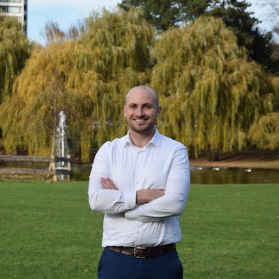 Ein junger Mann mit Glatze und Bart steht mit verschränkten Armen vor einem Teich in einem Park.
