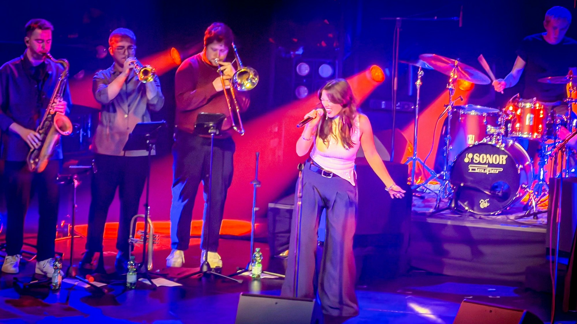 Die Leverkusener Sängerin Emily Otto steht auf einer Bühne und hält ein Mikrofon in Händen, links hinter ihr stehen drei Musiker, die Saxophon, Trompete und Posaune spielen, rechts steht ein Schlagzeug.