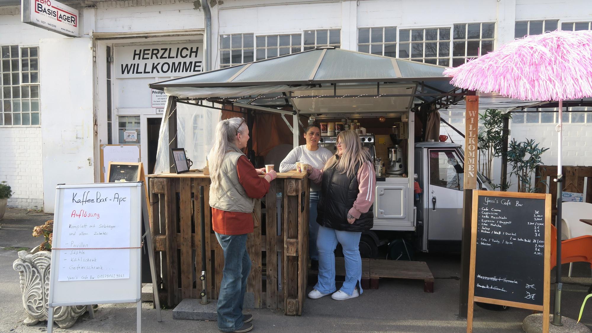 Drei Frauen stehen vor der hölzernen Theke eines Kaffeemobils, das vor einer großen Halle steht.
