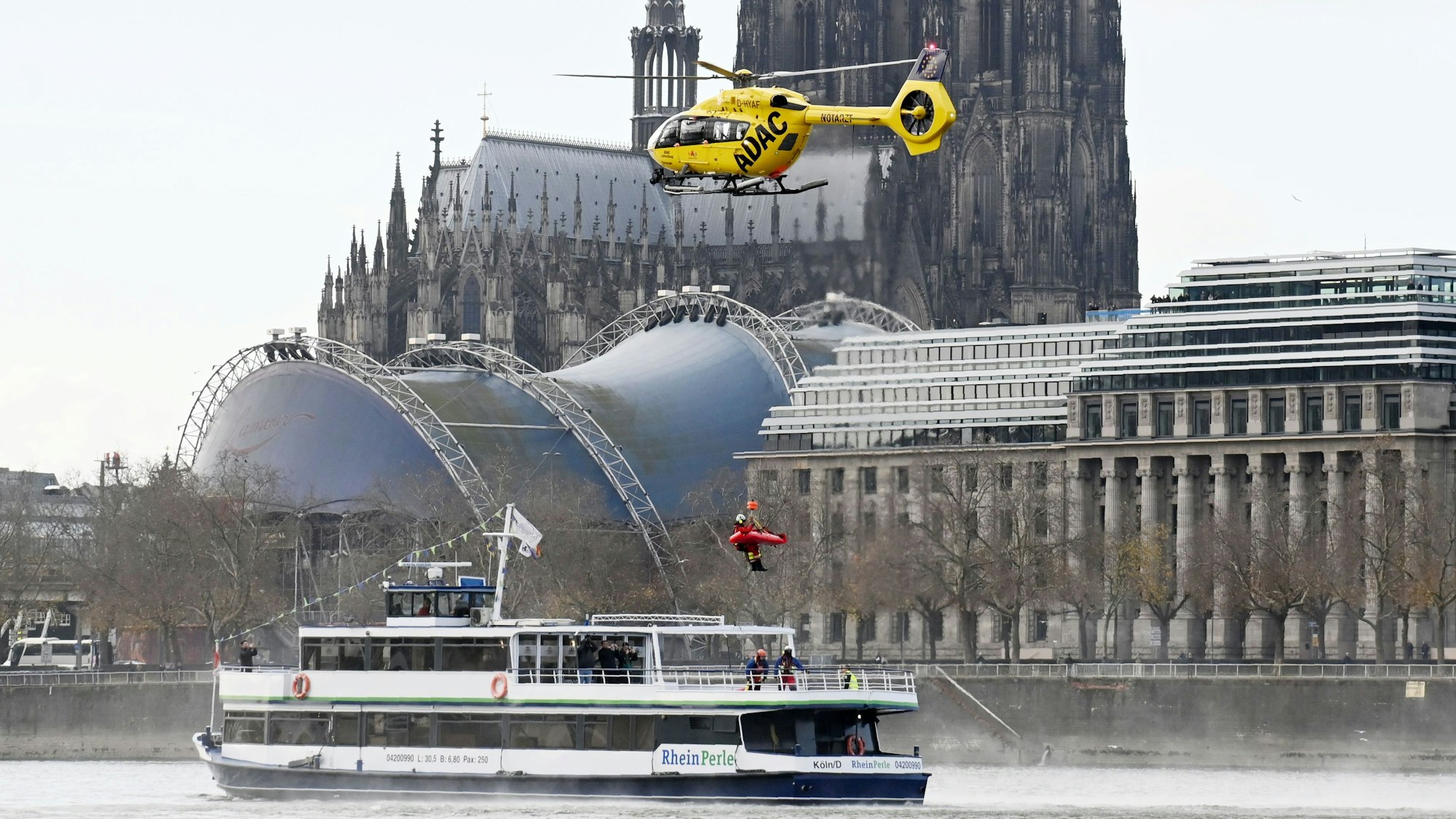 Ein ADAC-Hubschrauber schwebt auf dem Rhein vor dem Dom über einem Schiff, an seiner Winde hängt ein Retter mit einer Trage.