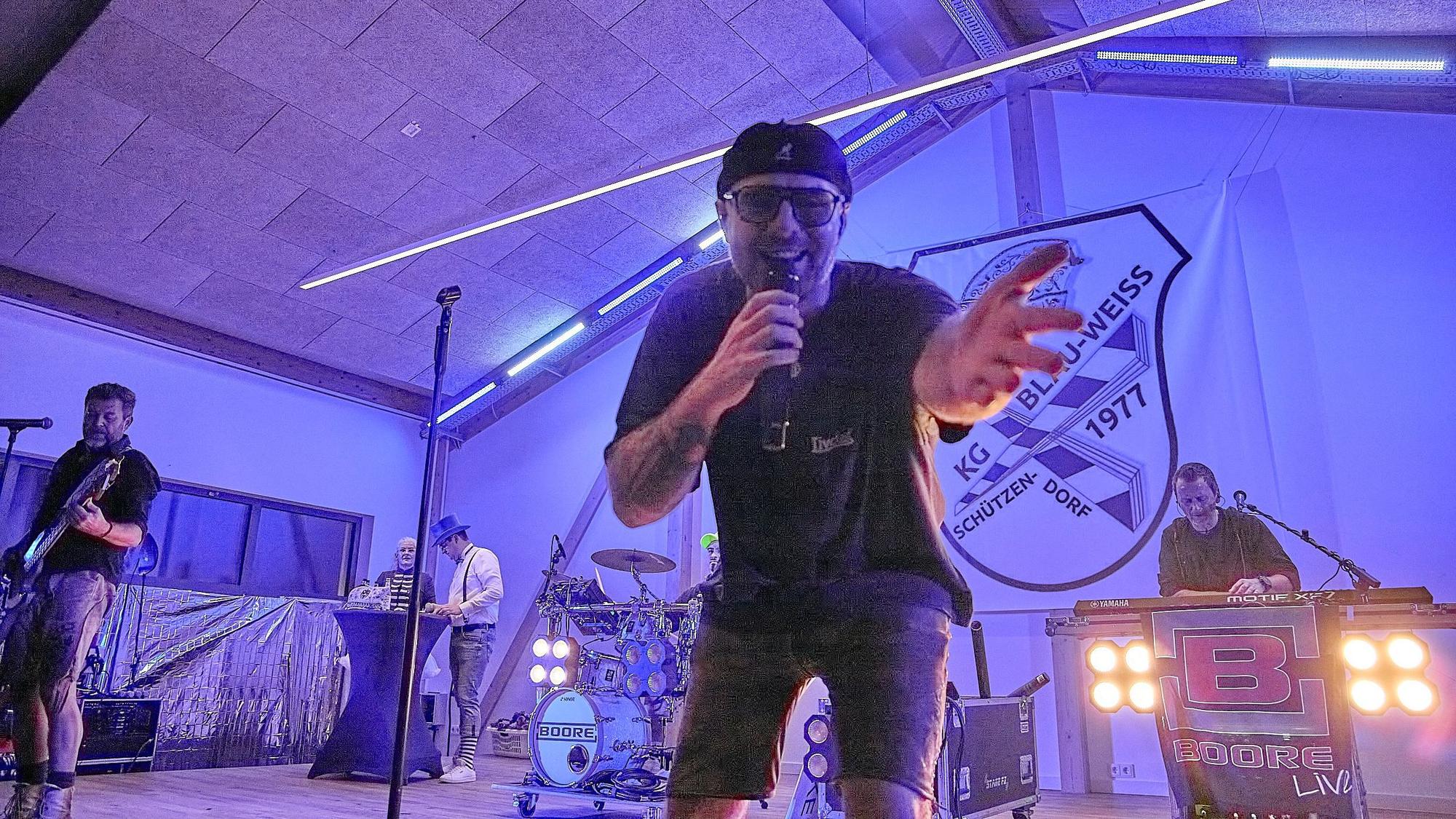 Der Frontmann der Boore beugt sich ins Mikrofon singend in Richtung Kamera, im Hintergrund sieht man die Bandkollegen musizieren.