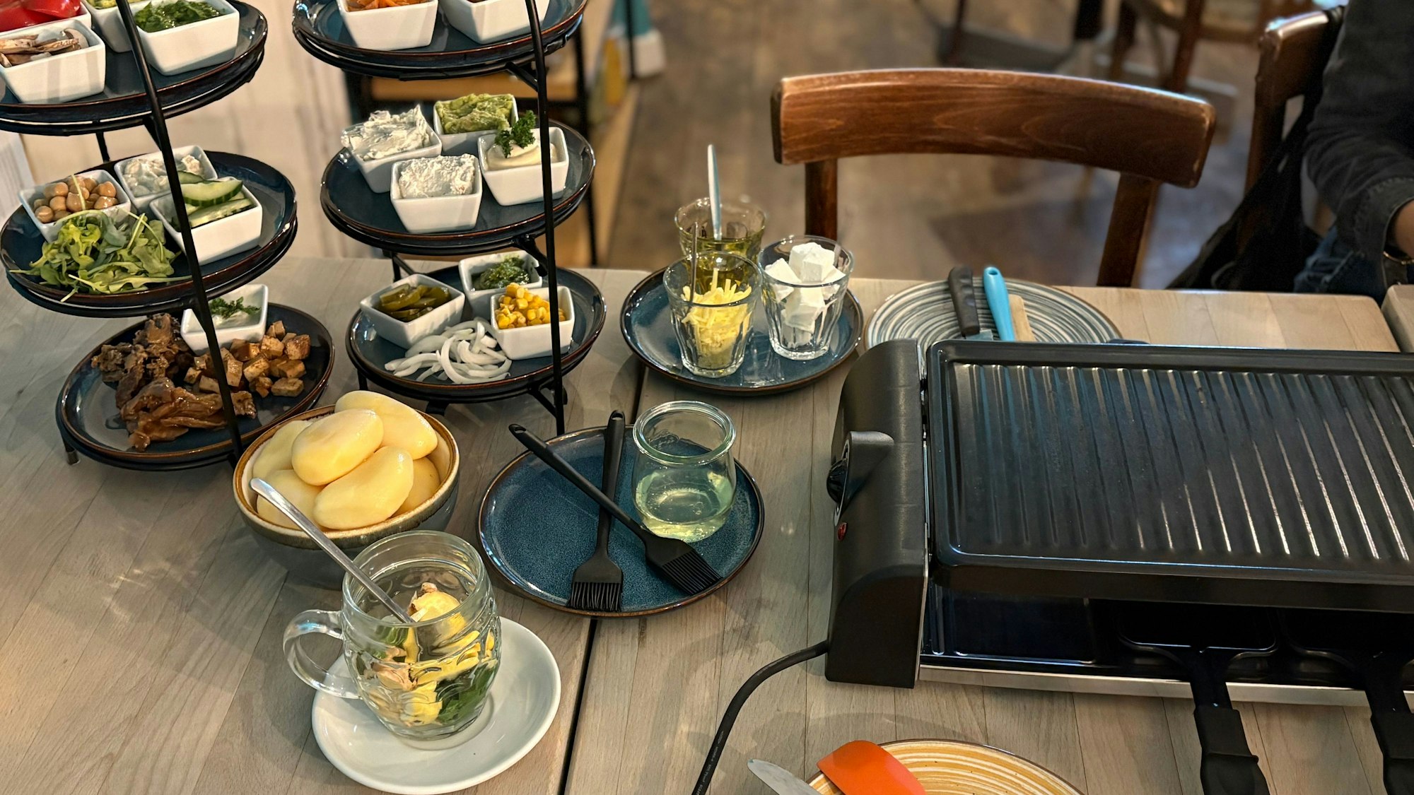 Ein Raclettegrill steht auf einem Tisch im Impact Café. Daneben zwei Etageren mit vielen Zutaten.