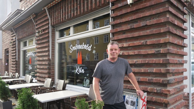 Ein Mann im grauen T-Shirt steht vor einem Lokal. Auch Tische und Stühle stehen vor der Fensterfront des Ladenlokals.