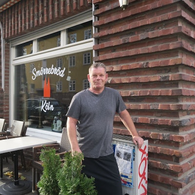 Ein Mann im grauen T-Shirt steht vor einem Lokal. Auch Tische und Stühle stehen vor der Fensterfront des Ladenlokals.
