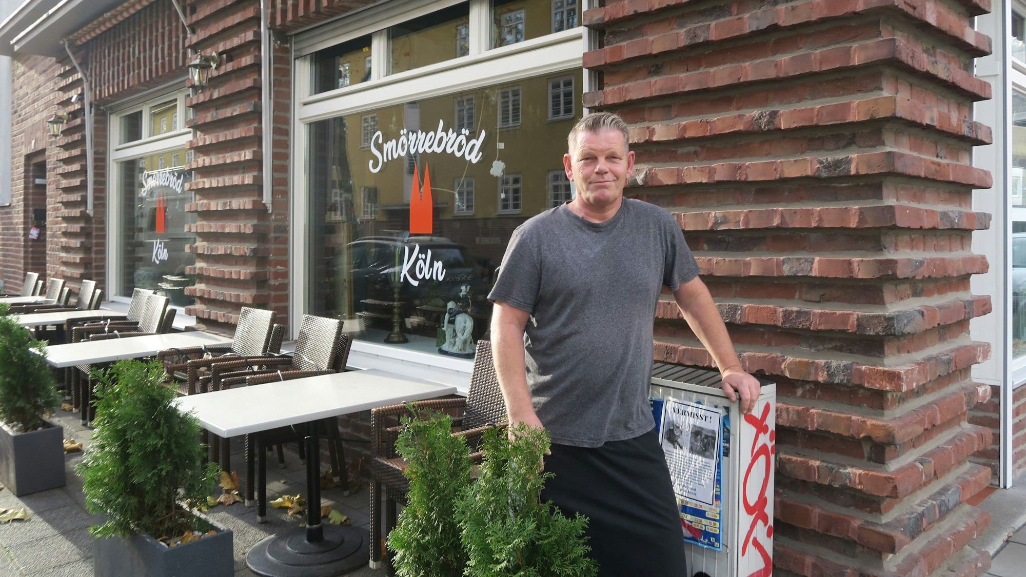 Ein Mann im grauen T-Shirt steht vor einem Lokal. Auch Tische und Stühle stehen vor der Fensterfront des Ladenlokals.