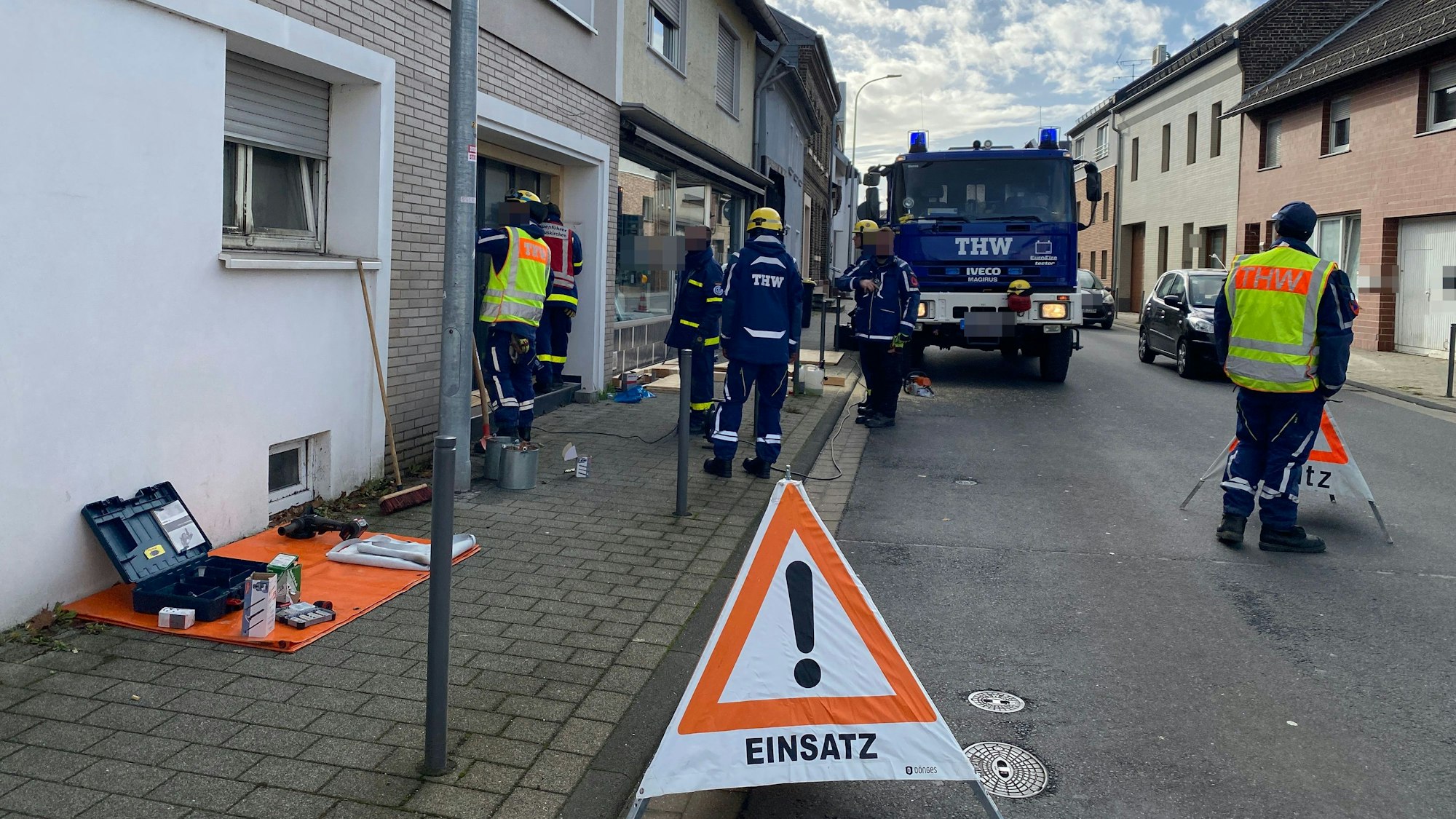Auf der Kölner Straße stehen Mitglieder der Feuerwehr und des THW, die an einem Hauseingang arbeiten. Auf der Straße steht ein Warndreieck mit der Aufschrift „Einsatz“.