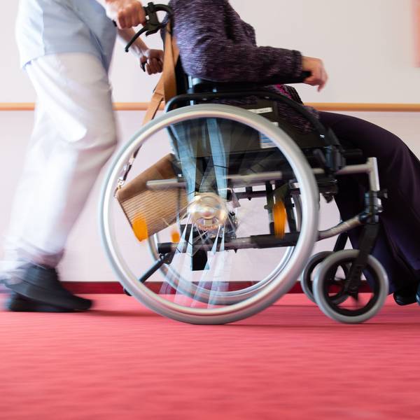 Ein Pfleger schiebt eine Bewohnerin eines Pflegeheims in einem Rollstuhl über den Flur.