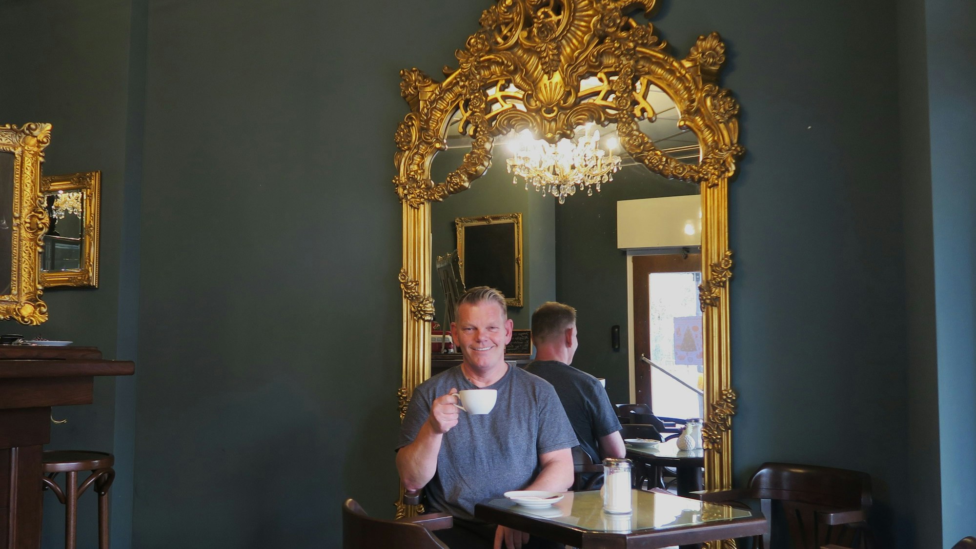 Ein Mann sitzt vor einem großen Spiegel mit Goldrand an einem Hochtisch und trinkt Kaffee.