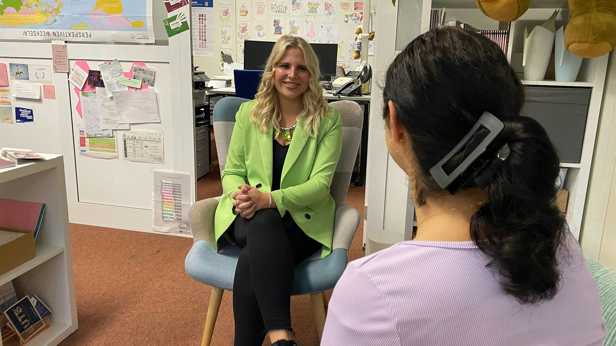 Eine Frau mit dunklen Haaren ist von hinten zu sehen. Sie redet mit einer blonden Frau, die gegenüber sitzt.