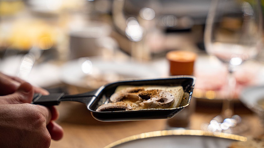 Ein Raclette-Pfännchen mit Käse und Pilzen wird gehalten, unscharf dahinter Teller und Gläser auf einem Tisch.