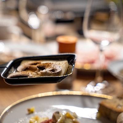 Ein Raclette-Pfännchen mit Käse und Pilzen wird gehalten, unscharf dahinter Teller und Gläser auf einem Tisch.