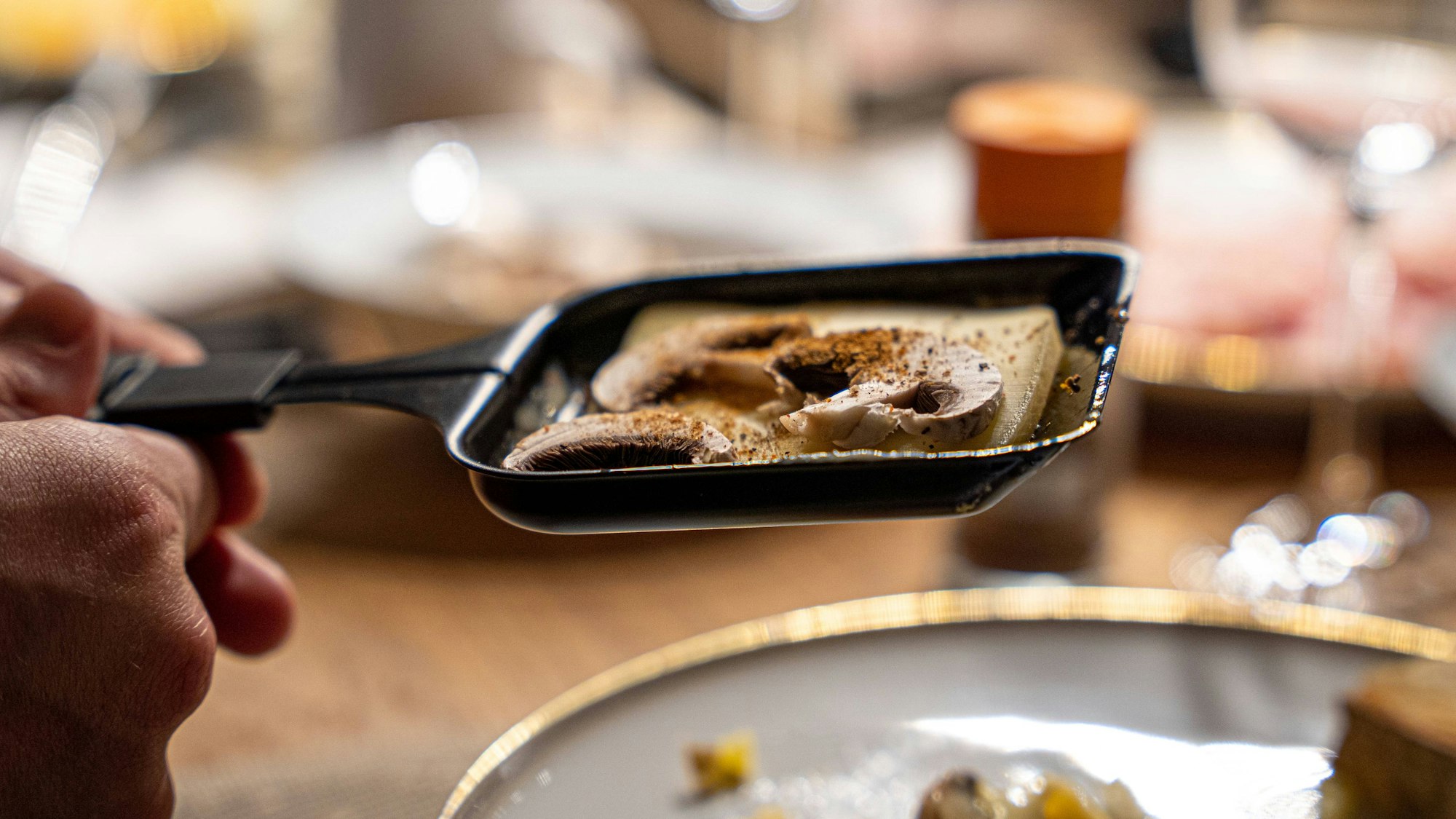 Ein Raclette-Pfännchen mit Käse und Pilzen wird gehalten, unscharf dahinter Teller und Gläser auf einem Tisch.