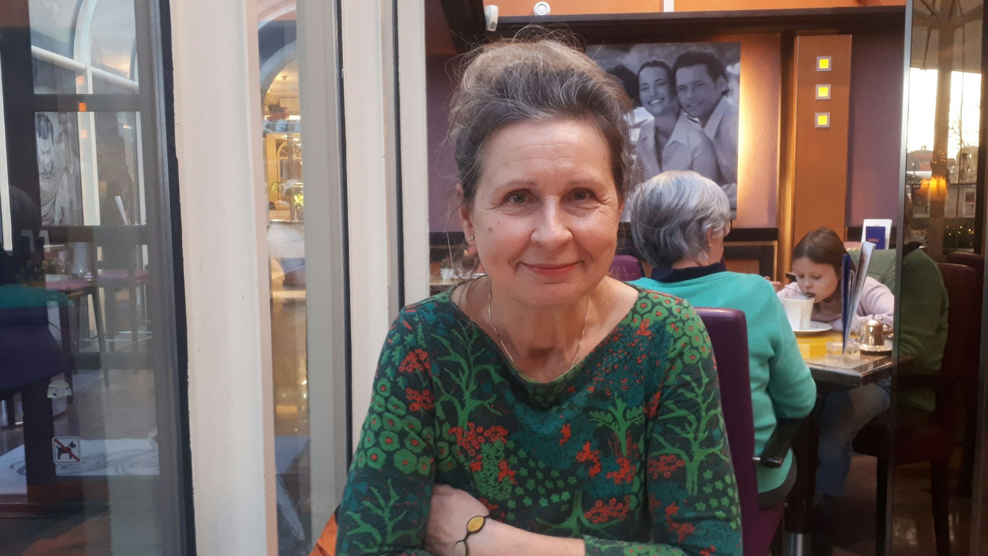 Eine Frau mit grün-rot geblümten Oberteil sitzt lächelnd und mit verschränkten Armen in einem Café.