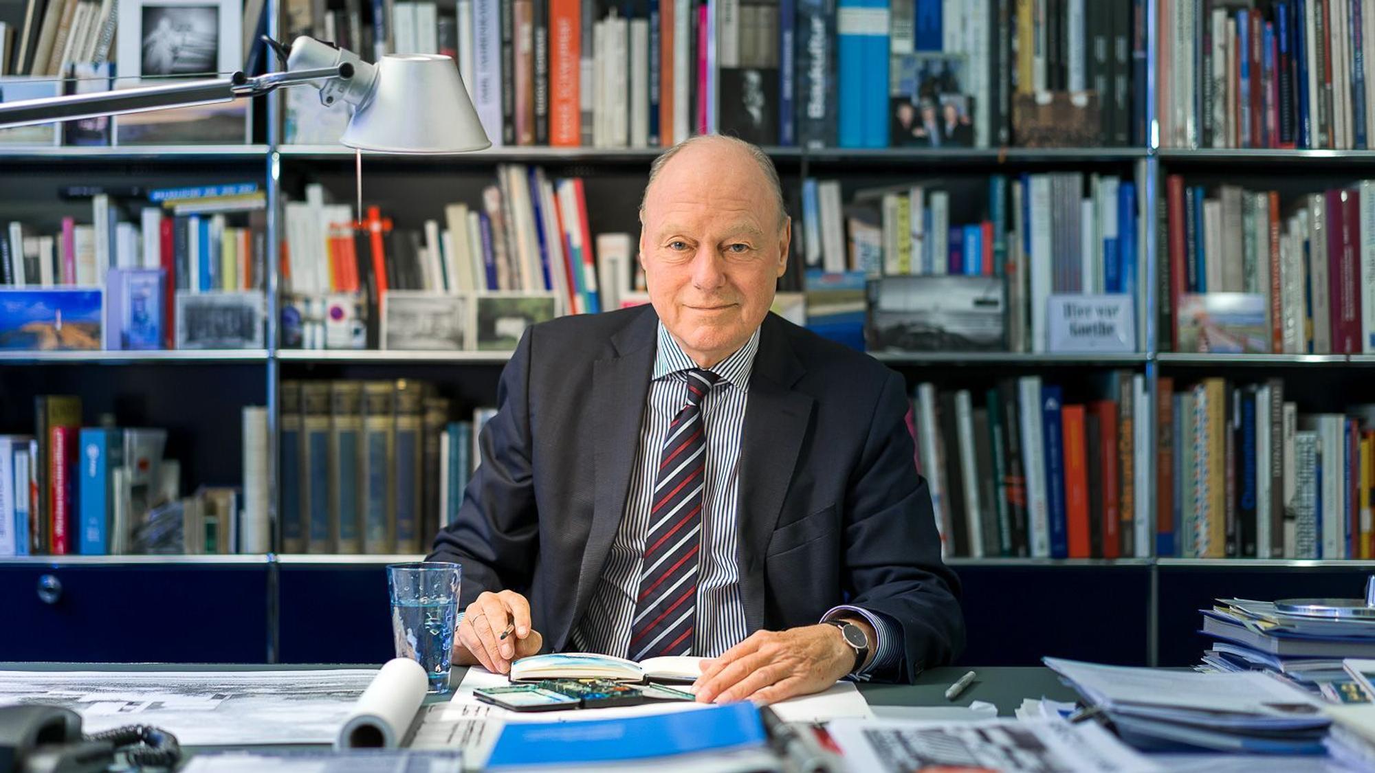 Kaspar Kraemer sitzt an einem vollen Schreibtisch und lächelt in die Kamera. Hinter ihm ein volles Bücherregal