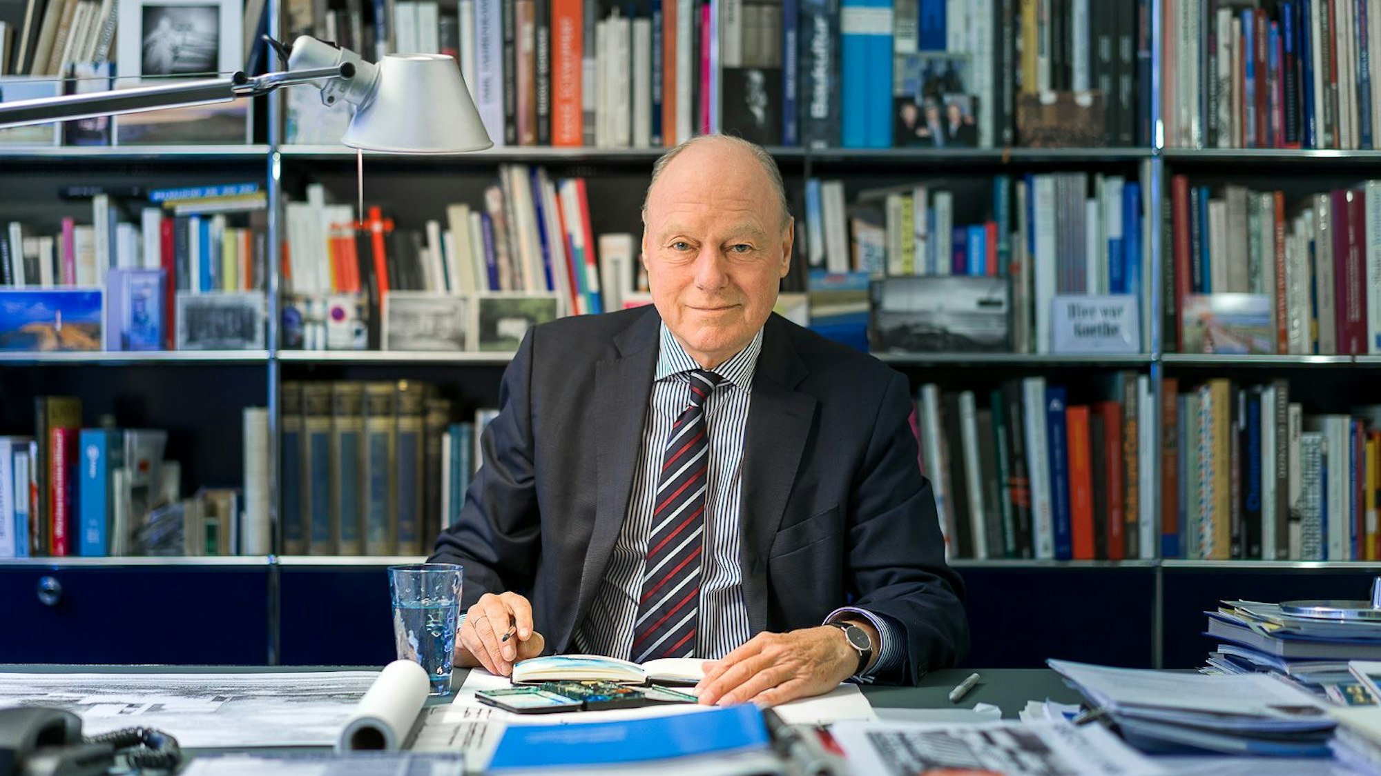 Kaspar Kraemer sitzt an einem vollen Schreibtisch und lächelt in die Kamera. Hinter ihm ein volles Bücherregal